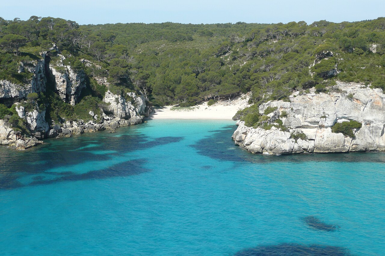 Sunlit white cliffs and a sandy cove above clear turquoise water at Cala Macarelleta, Menorca